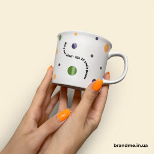 Біла чашка з друком у вигляді різнокольорових точок та тексту